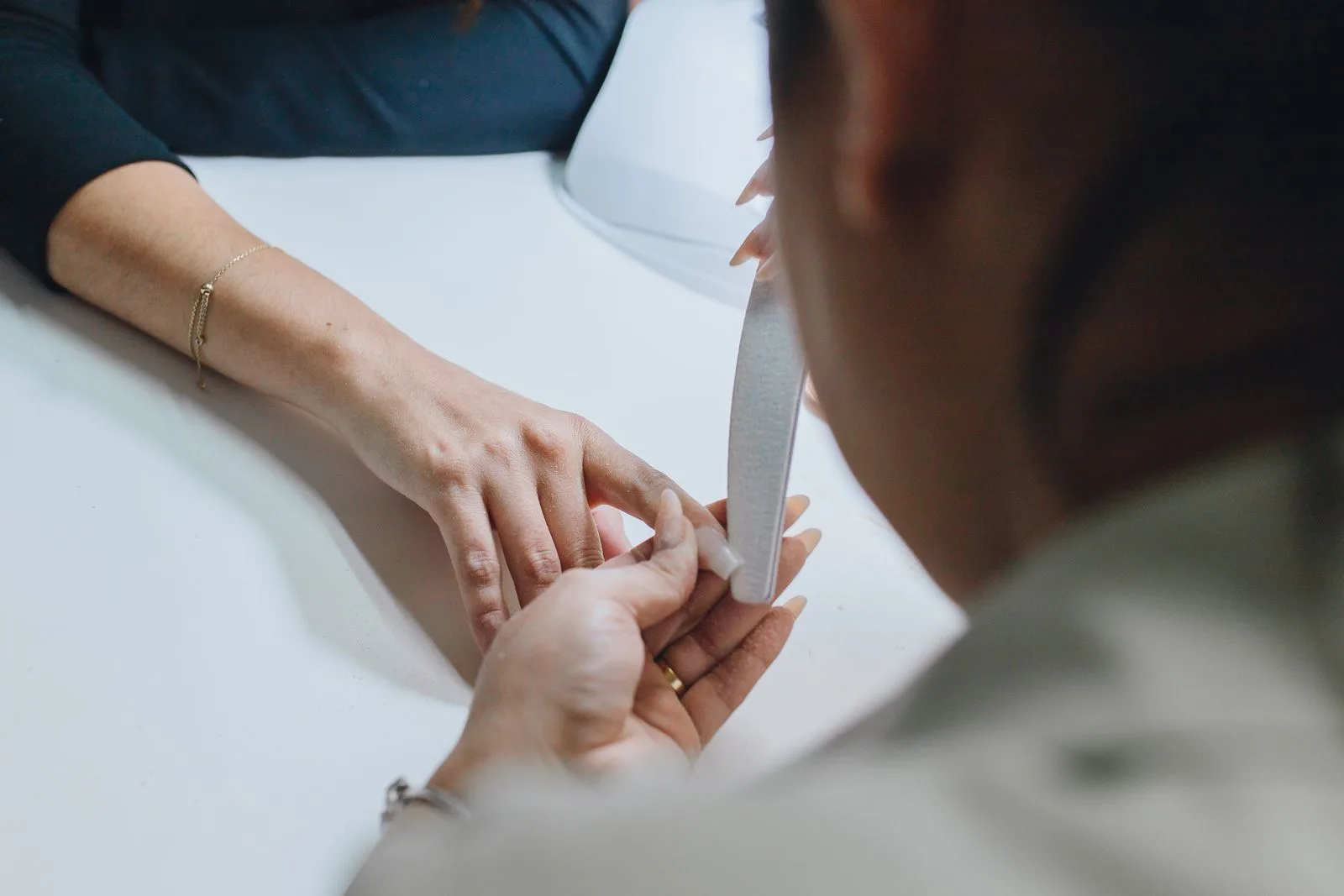 Tehniciană pilind unghiile unei cliente, văzută de deasupra mesei